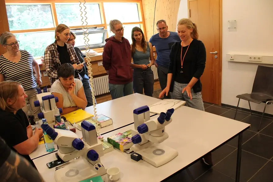 Die Vorstellung der Heimarbeiten im Kursraum machte allen Beteiligten viel Spaß (Foto: Thomas Hövelmann) Eine Teilnehmerin präsentiert ihre Ergebnisse vor einer kleinen Gruppe an einem Tisch mit Mikroskopen und Bestimmungsmaterial.